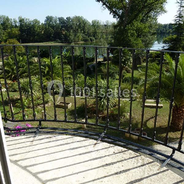 Vue depuis un balcon avec une rambarde en fer forgé, donnant sur un jardin verdoyant et un plan d'eau en arrière-plan.