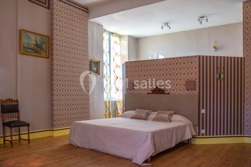 Chambre lumineuse avec un lit double, parquet au sol, murs décorés de motifs et une grande fenêtre avec rideaux.