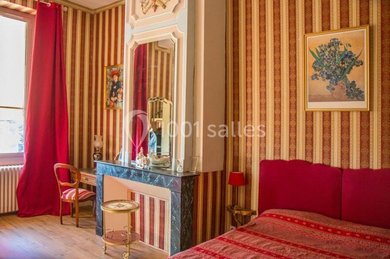 Chambre avec lit rouge, cheminée en marbre, miroir, fauteuil en bois et murs ornés de rayures rouges et dorées.