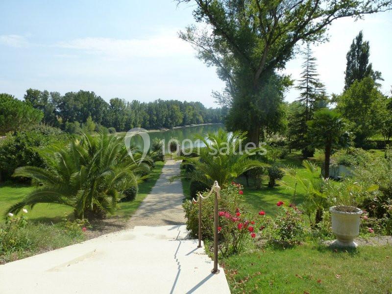 Vue sur un jardin verdoyant avec palmiers, rosiers et un chemin menant à un lac entouré d'arbres.