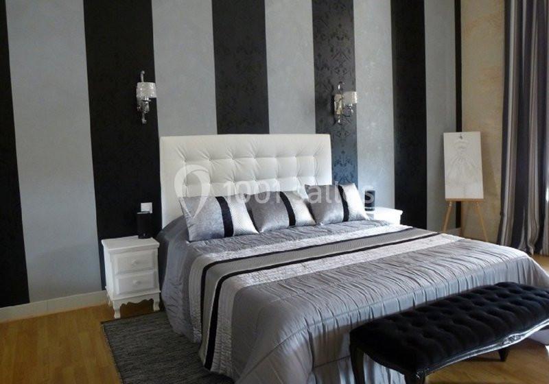 Lit double avec tête de lit capitonnée, draps gris, coussins décoratifs, et murs rayés noir et gris.