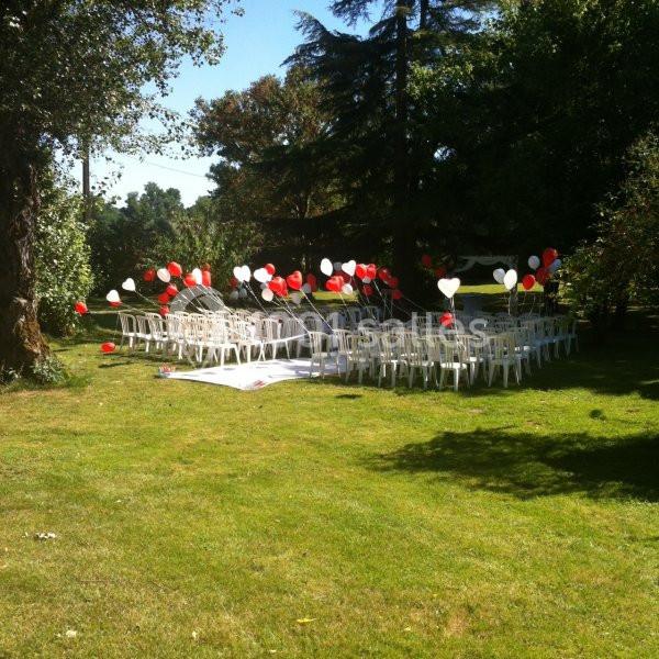 Chaises blanches disposées en extérieur sur une pelouse, décorées de ballons rouges et blancs, sous des arbres.