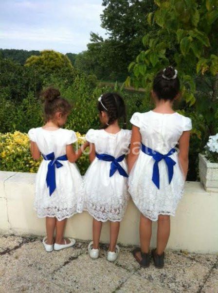 Trois jeunes filles en robe blanche avec des ceintures bleues, de dos, regardant un jardin verdoyant.
