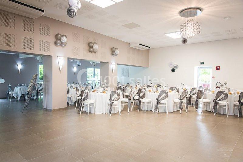 Salle de réception lumineuse avec tables rondes dressées, décorations grises et blanches, et miroirs sur un mur.