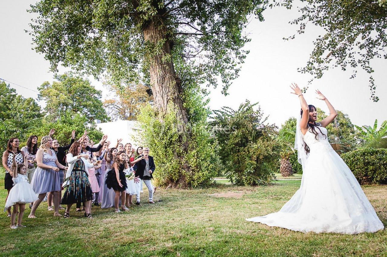 Une mariée lance son bouquet à un groupe de femmes rassemblées dans un jardin lors d'une réception en plein air.