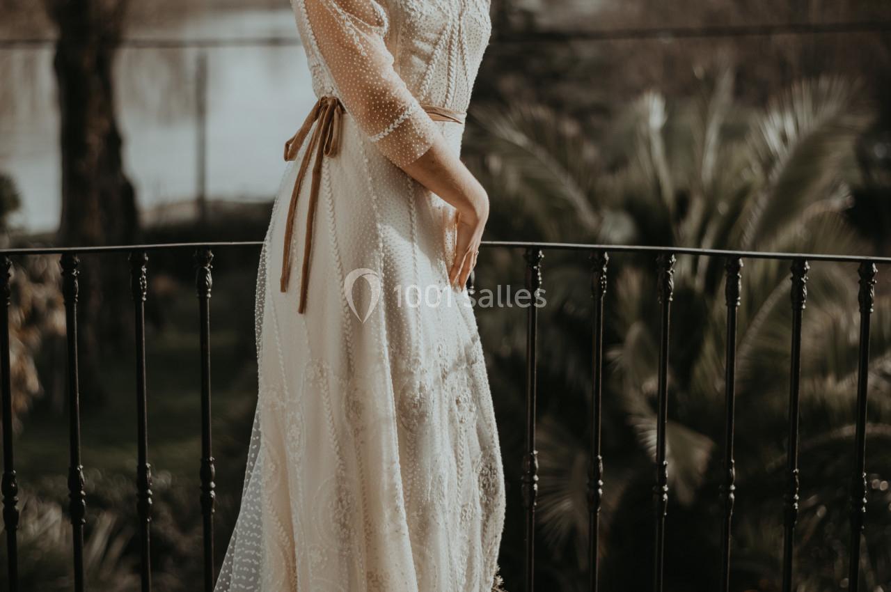 Femme en robe longue en dentelle blanche posant sur un balcon avec un paysage naturel en arrière-plan.