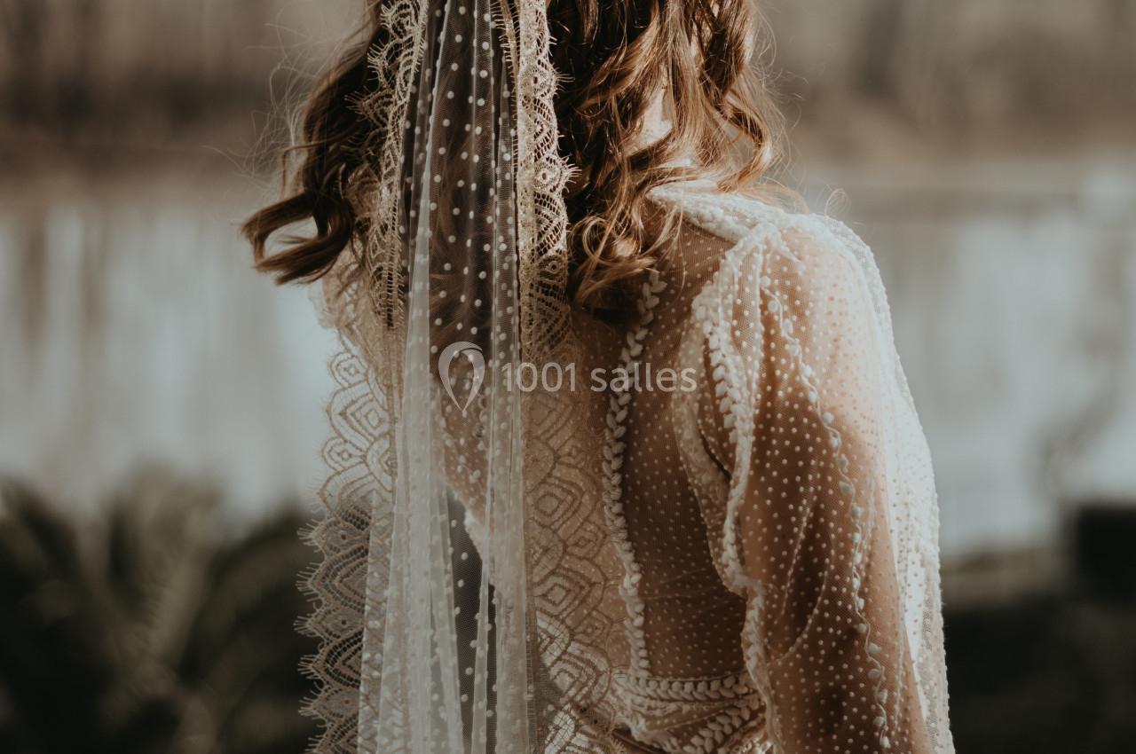 Femme de dos portant une robe en dentelle et un voile, debout sur un balcon avec un paysage fluvial en arrière-plan.