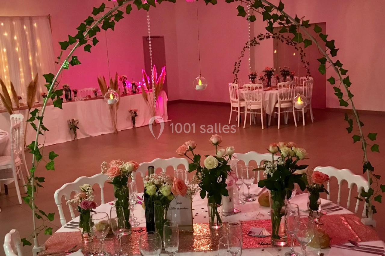 Table de réception décorée avec des fleurs, nappes blanches, et guirlandes lumineuses dans une salle éclairée en rose.