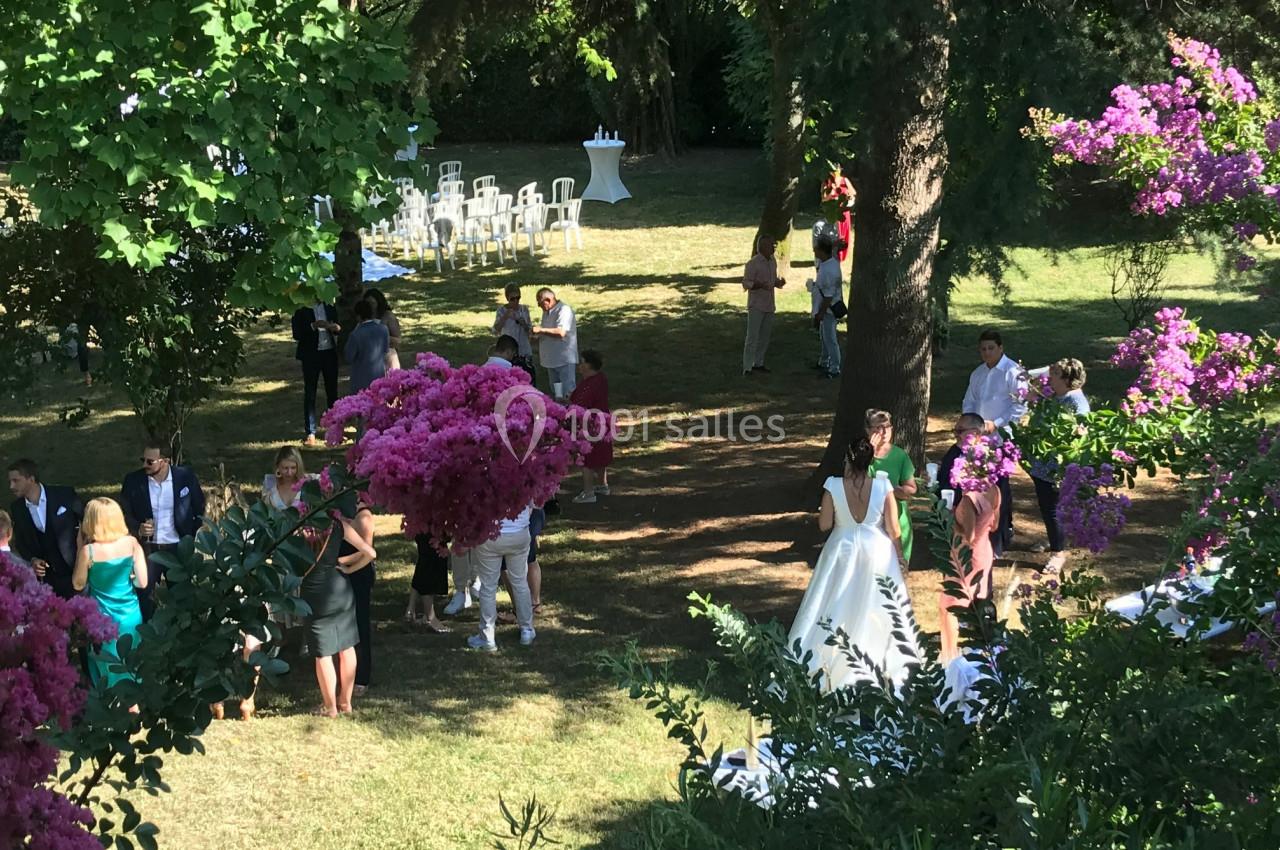 Des invités discutent dans un jardin ombragé lors d'une réception, avec des fleurs violettes et des chaises blanches en…