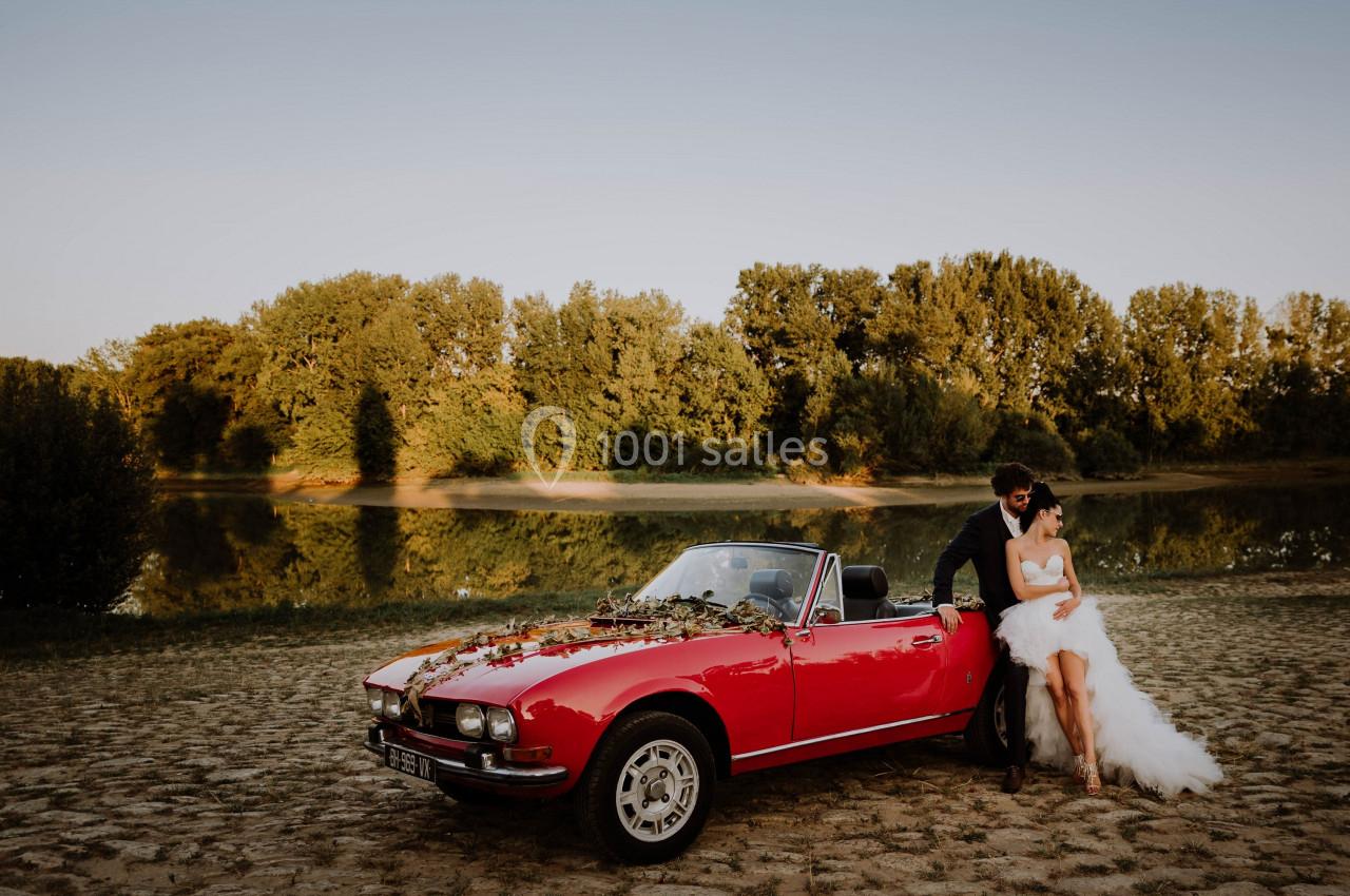 Un couple en tenue de mariage pose près d'une voiture décapotable rouge ornée de fleurs, devant un paysage naturel.