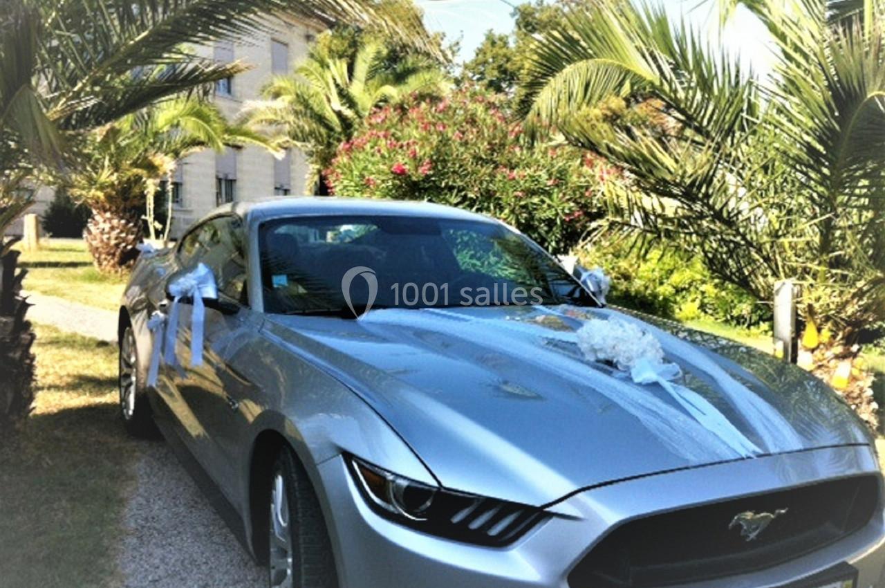 Voiture argentée décorée pour un mariage, stationnée sur une allée bordée de palmiers devant un bâtiment clair.