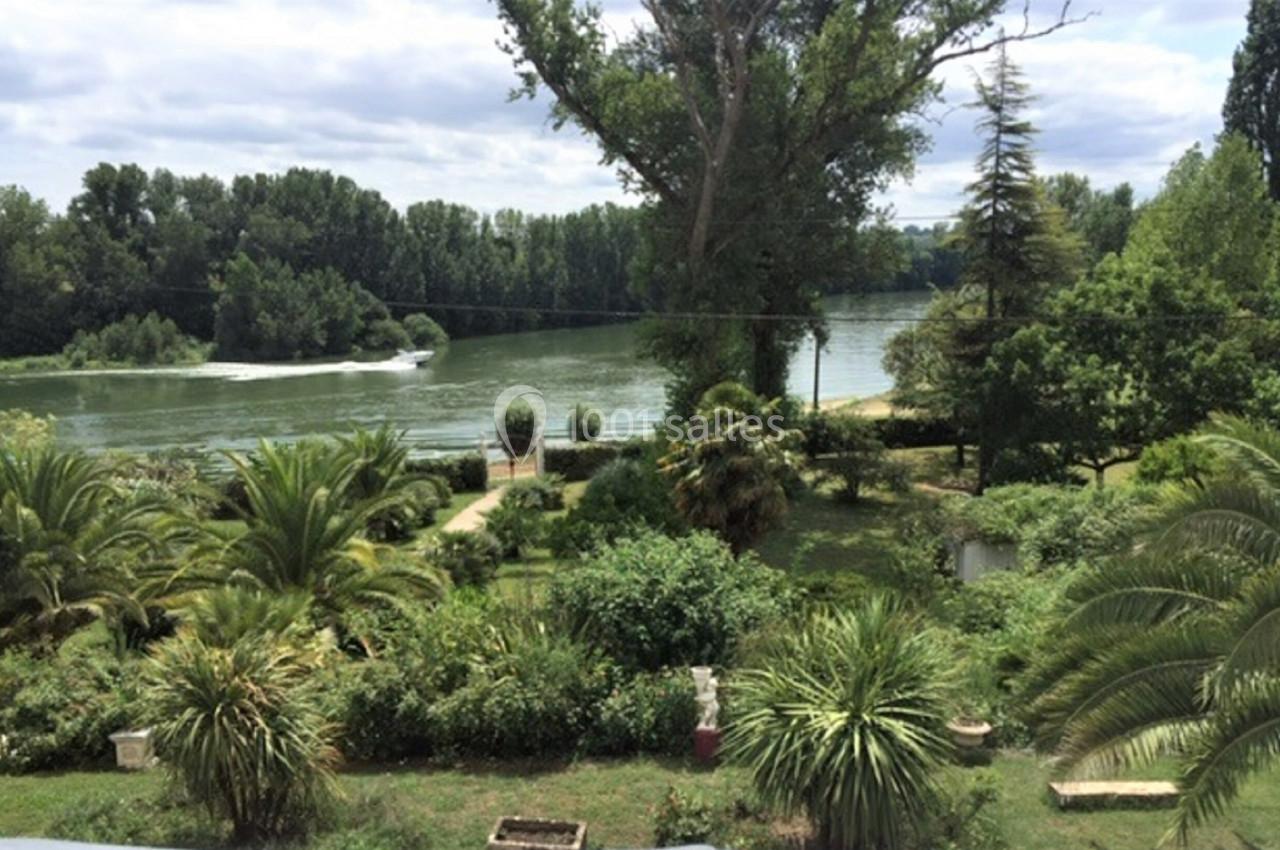 Jardin verdoyant avec des palmiers et des arbres, bordé par une rivière sous un ciel partiellement nuageux.