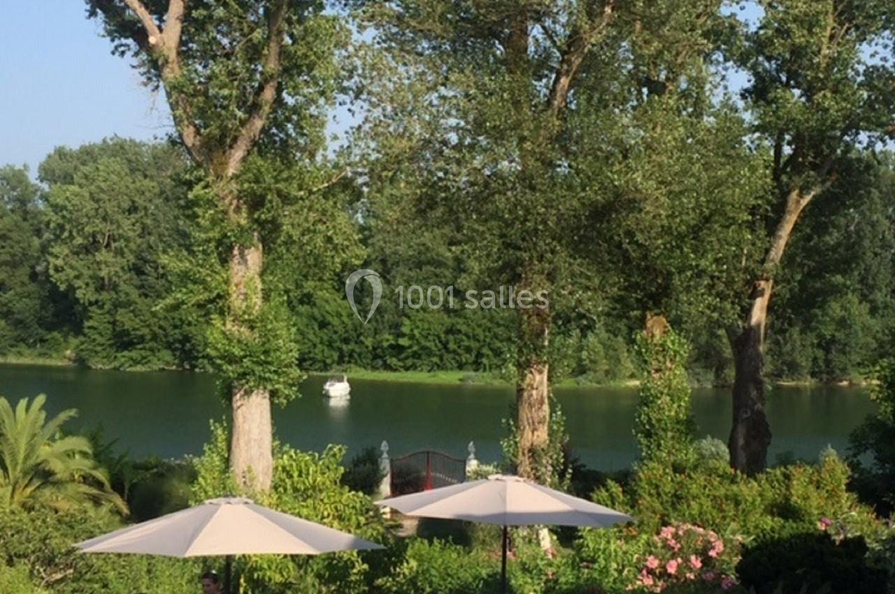 Vue sur un jardin verdoyant avec parasols, bordé par une rivière calme et entouré d'arbres en été.