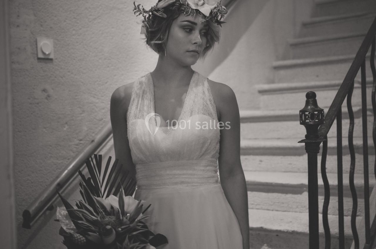Une femme en robe de mariée tient un bouquet tropical, debout près d'un escalier en intérieur.