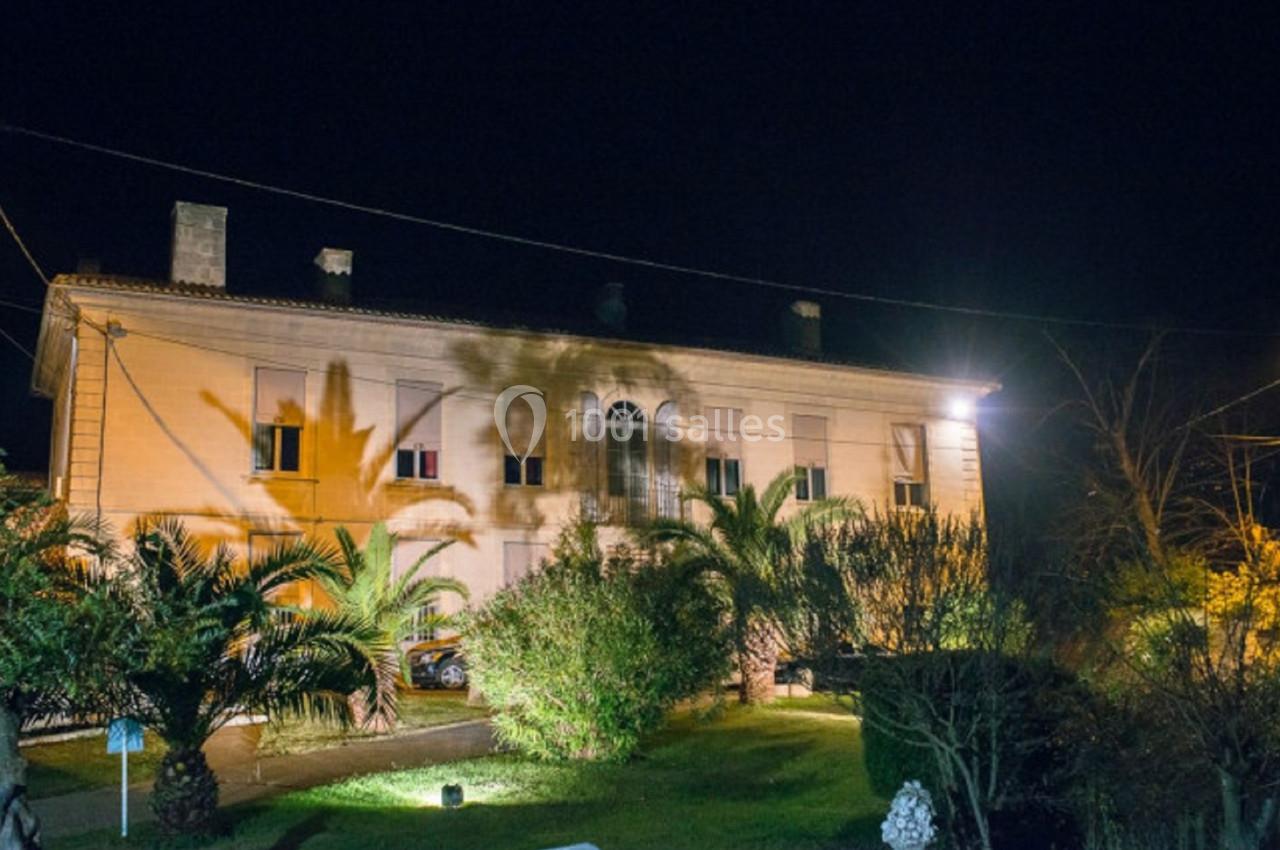 Façade d'une grande maison éclairée la nuit, entourée de palmiers et d'un jardin.