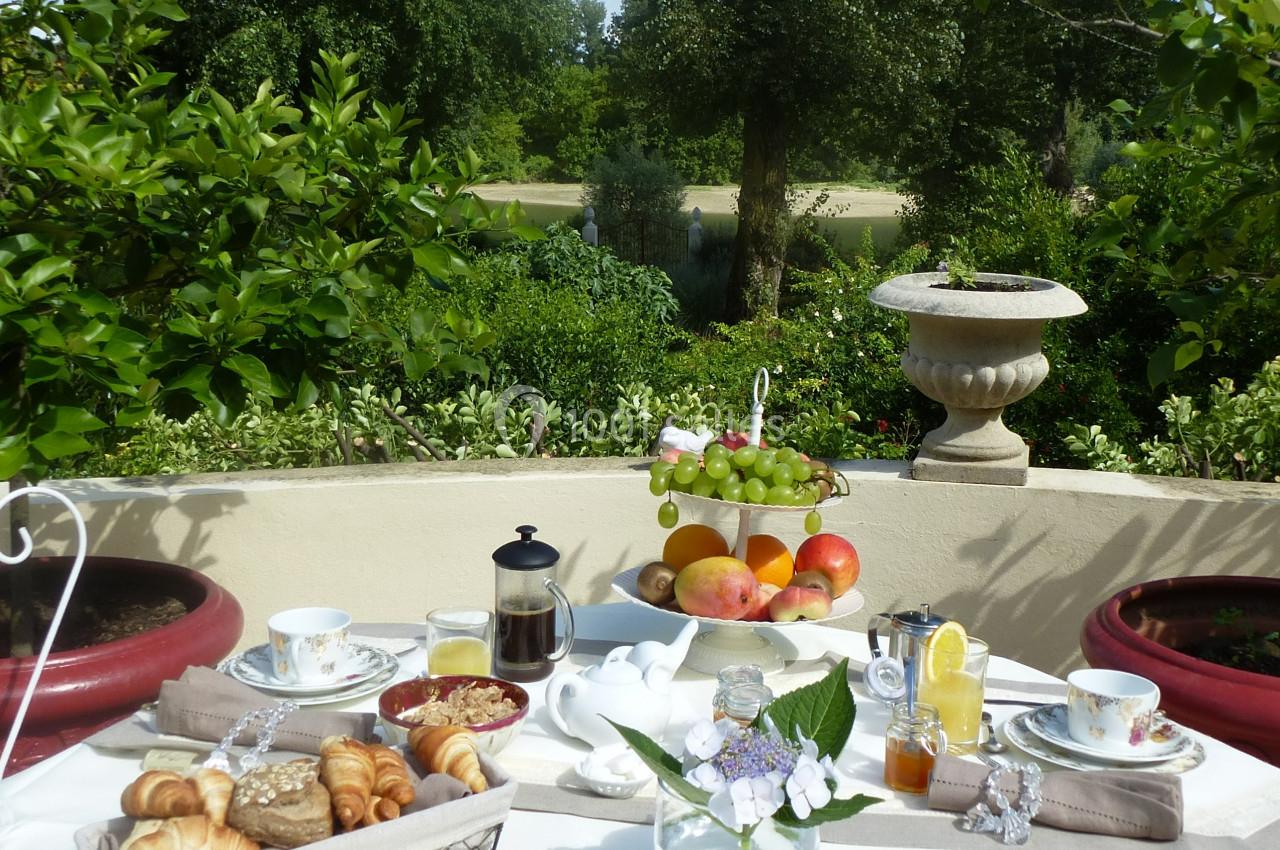 Petit-déjeuner en plein air sur une table dressée avec viennoiseries, fruits et boissons, entourée de verdure.