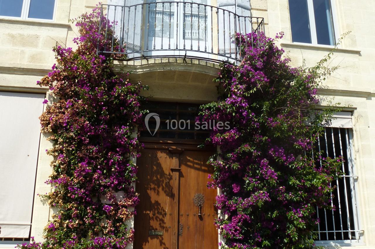Façade en pierre avec une porte en bois encadrée de bougainvilliers fleuris et un balcon avec volets ouverts.