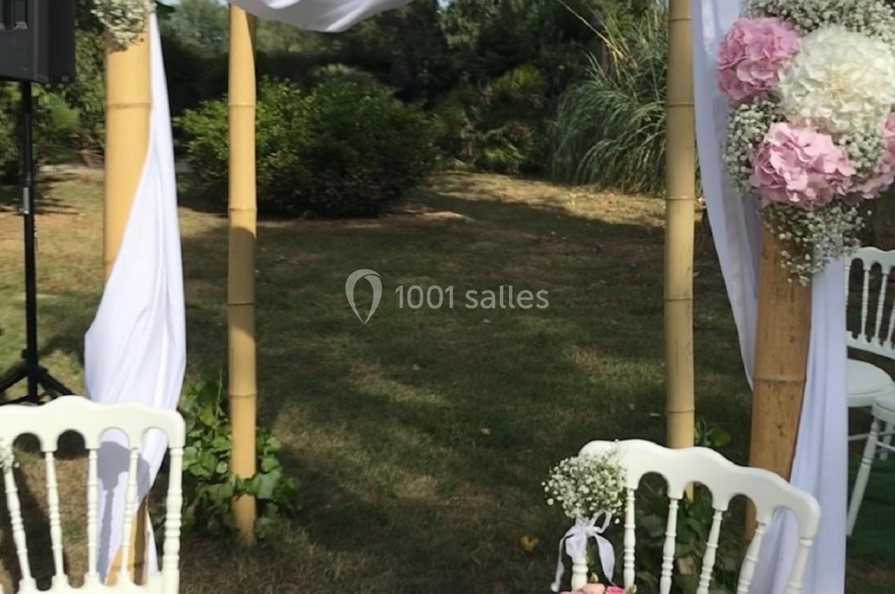 Arche de mariage en bambou décorée de fleurs roses et blanches, entourée de chaises blanches dans un jardin.