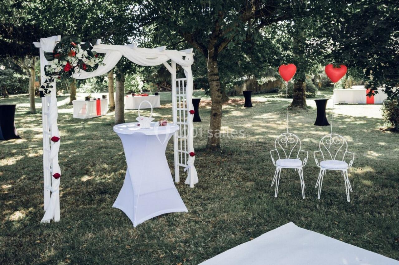 Arche décorée de fleurs blanches et rouges, table haute et chaises blanches avec ballons en forme de cœur dans un jardin.
