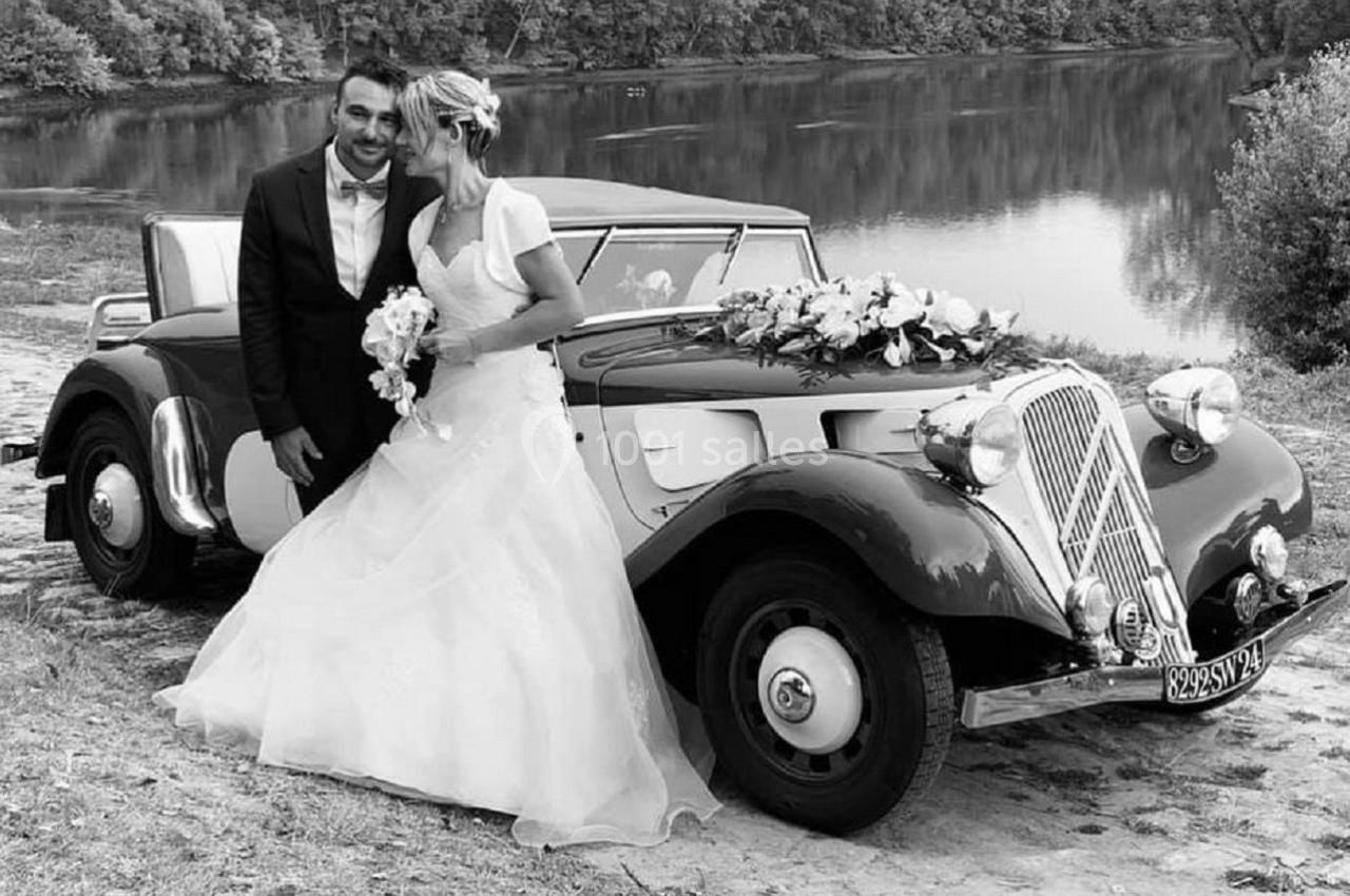 Un couple en tenue de mariage pose devant une voiture ancienne décorée de fleurs, près d'un lac et d'arbres.