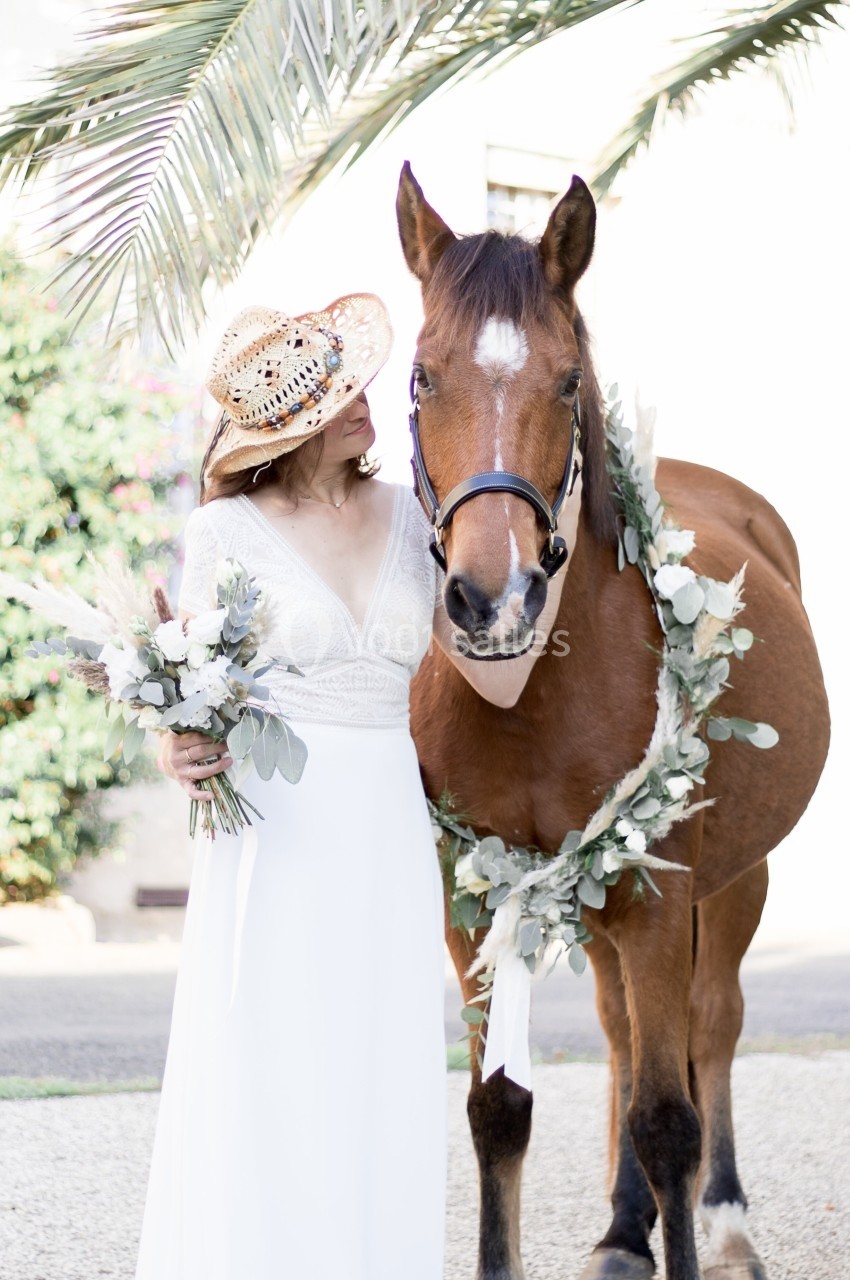 Femme en robe blanche tenant un bouquet, debout à côté d'un cheval décoré d'une guirlande florale.