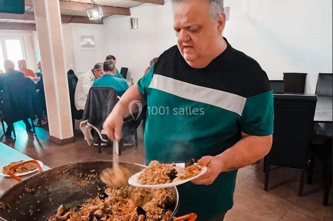 Un homme sert de la paella avec une louche dans une assiette, dans une salle avec des convives assis.