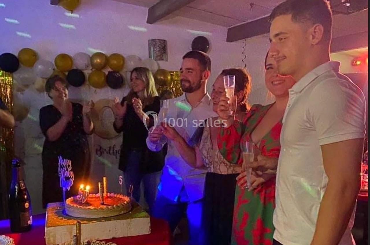 Des personnes réunies autour d'un gâteau d'anniversaire, tenant des verres, dans une pièce décorée de ballons et guirlandes.