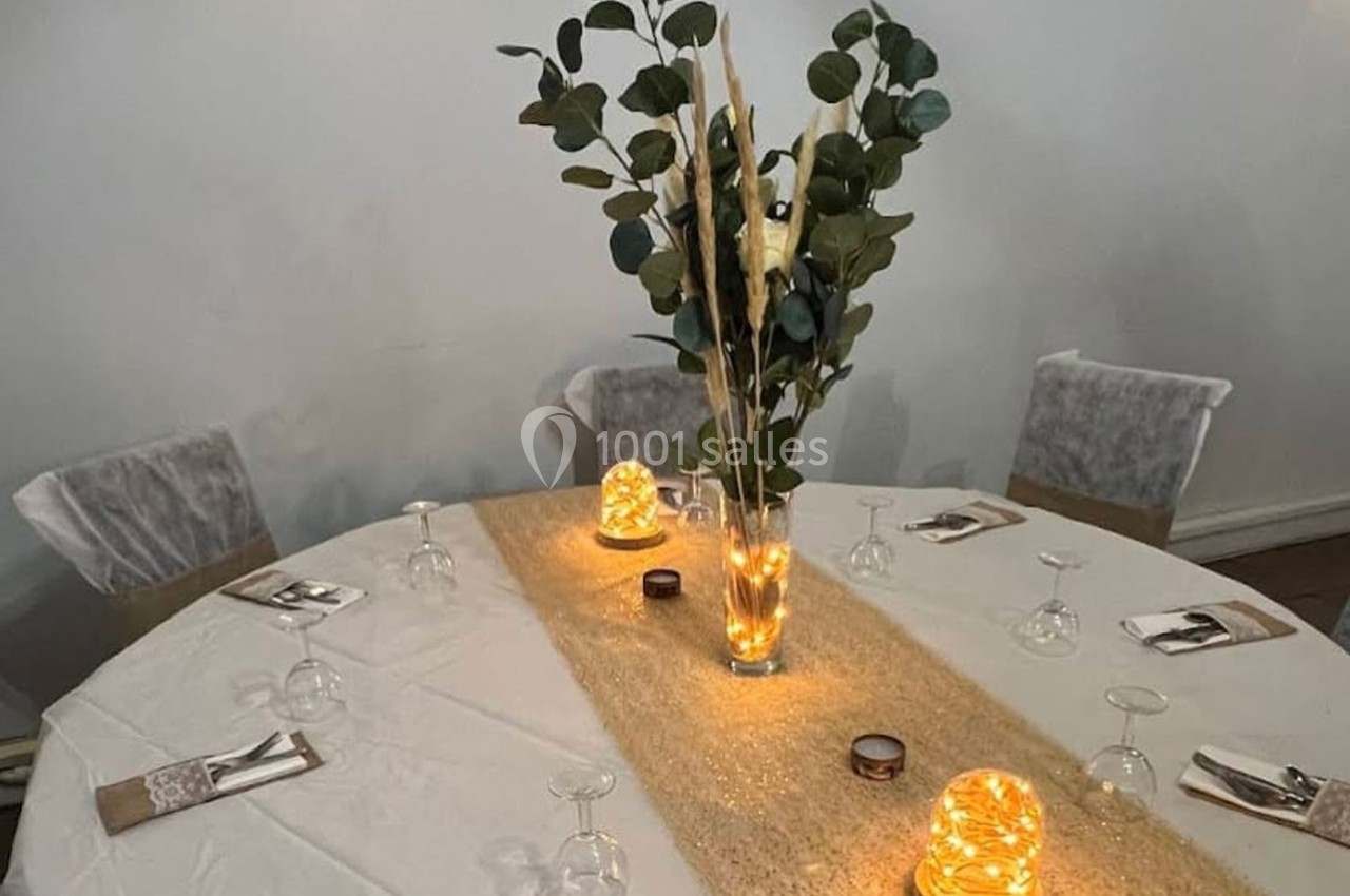 Table ronde décorée avec une nappe blanche, un chemin de table doré, des bougies lumineuses et un vase de feuillage.