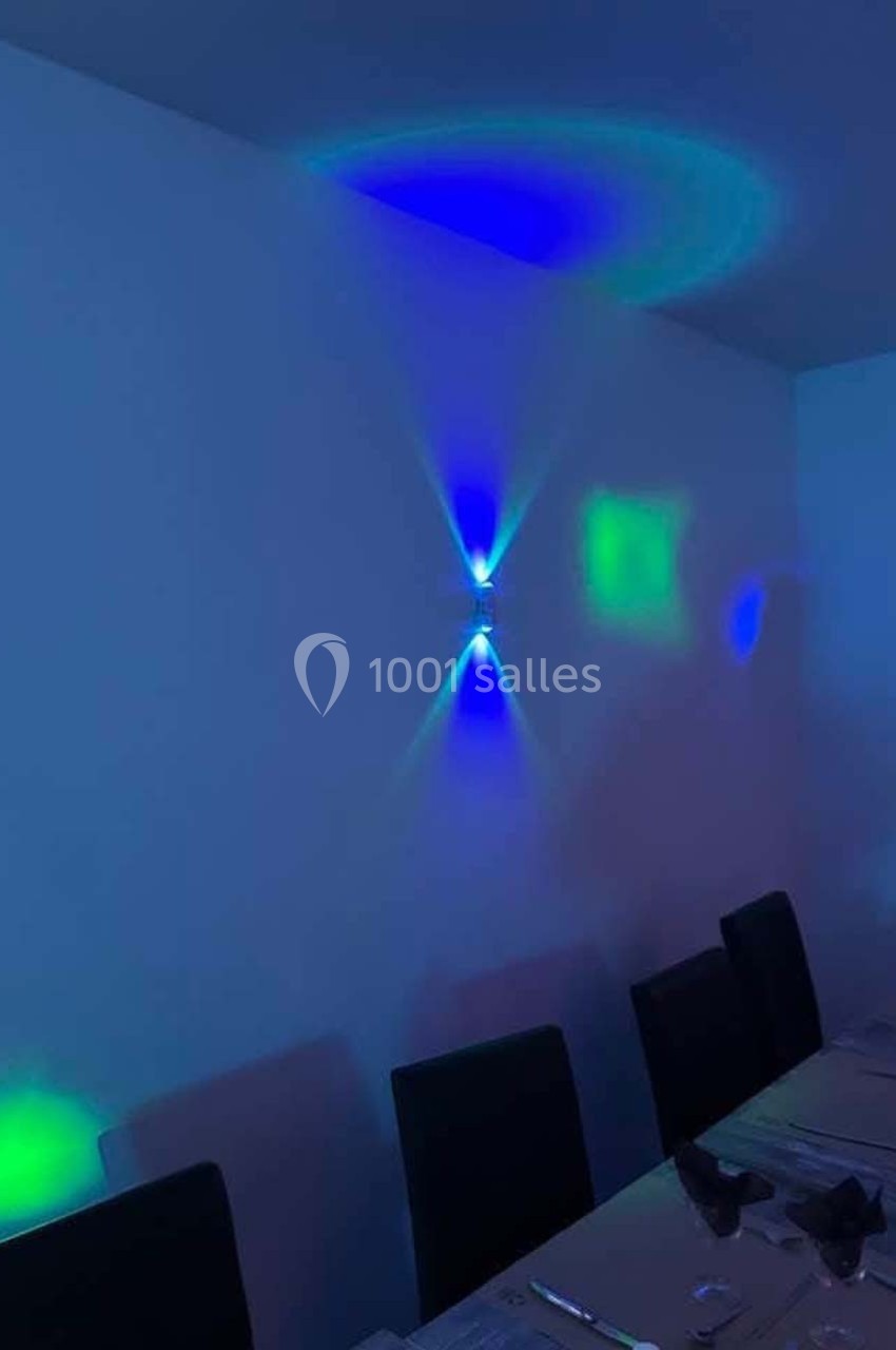 Éclairage mural diffusant des lumières bleues et vertes dans une salle avec une table dressée et des chaises noires.