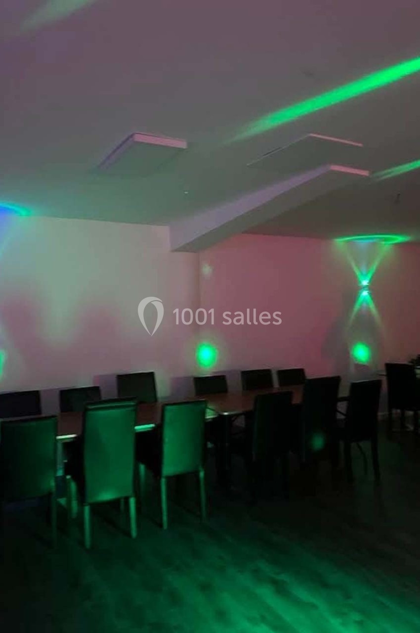 Salle sombre avec des tables alignées, des chaises noires et des lumières vertes et bleues projetées sur les murs.