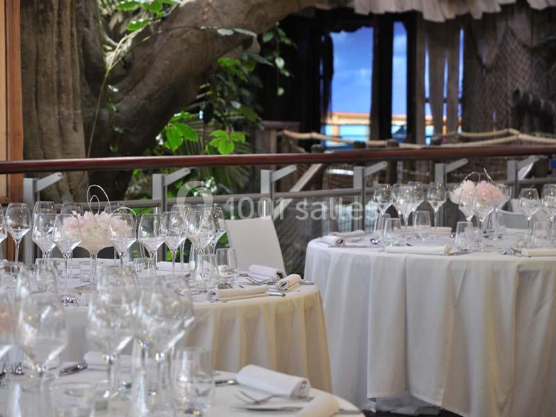 Salle de restaurant élégante avec tables rondes dressées, nappes blanches, verres à pied et décor végétal en arrière-plan.