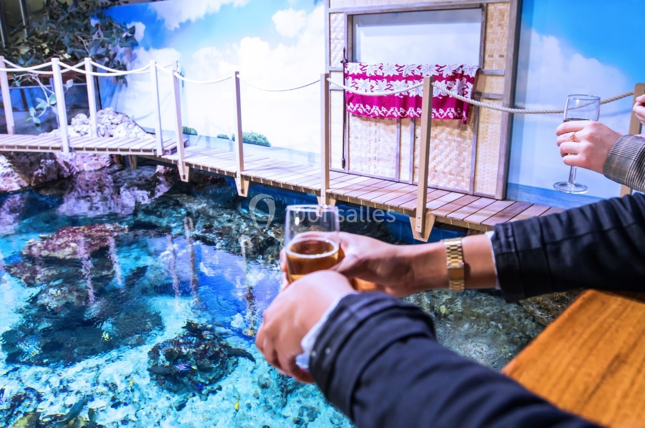Location salle Montpellier (Hérault) - Planet Ocean Montpellier #5 Deux personnes tenant des verres devant un bassin d'eau claire avec un pont en bois et une décoration murale.