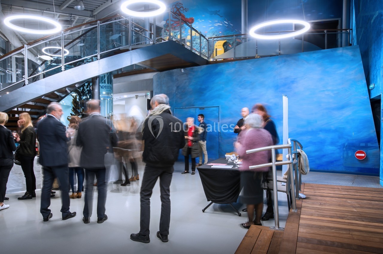 Des personnes discutent dans un hall moderne avec un mur bleu, un escalier en colimaçon et des éclairages circulaires.