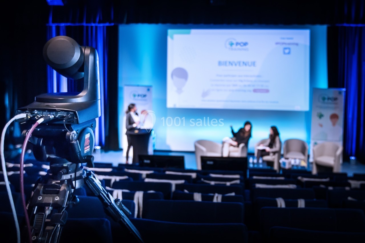 Caméra filmant une scène avec deux personnes assises et une présentatrice debout dans une salle de conférence.