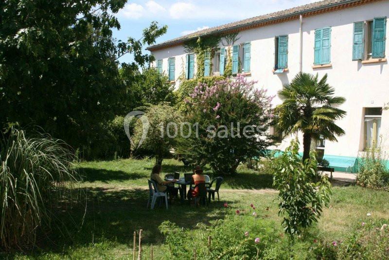 Trois personnes assises autour d'une table dans un jardin verdoyant devant une grande maison blanche aux volets bleus.