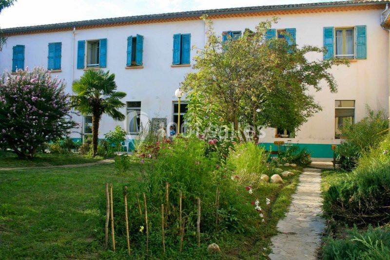 Façade d'un bâtiment à deux étages avec volets bleus, entouré d'un jardin fleuri et d'un chemin en pierre.