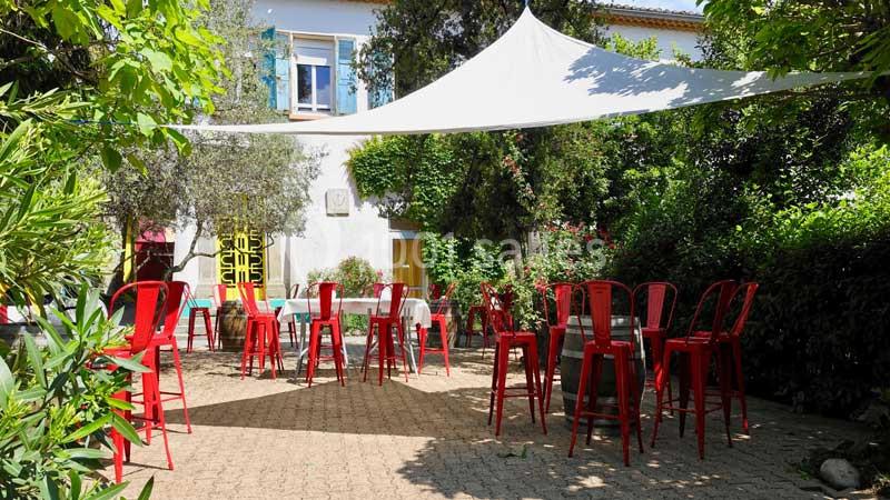 Terrasse extérieure avec des chaises rouges, tables et voile d'ombrage, entourée de végétation et d'un bâtiment blanc.