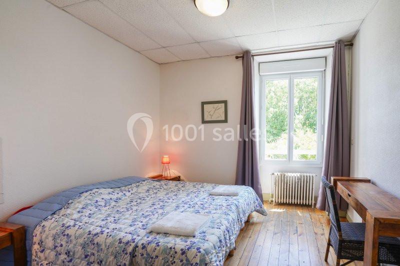 Chambre lumineuse avec lit double, couvre-lit fleuri, table de chevet avec lampe, fenêtre donnant sur un extérieur verdoyant.