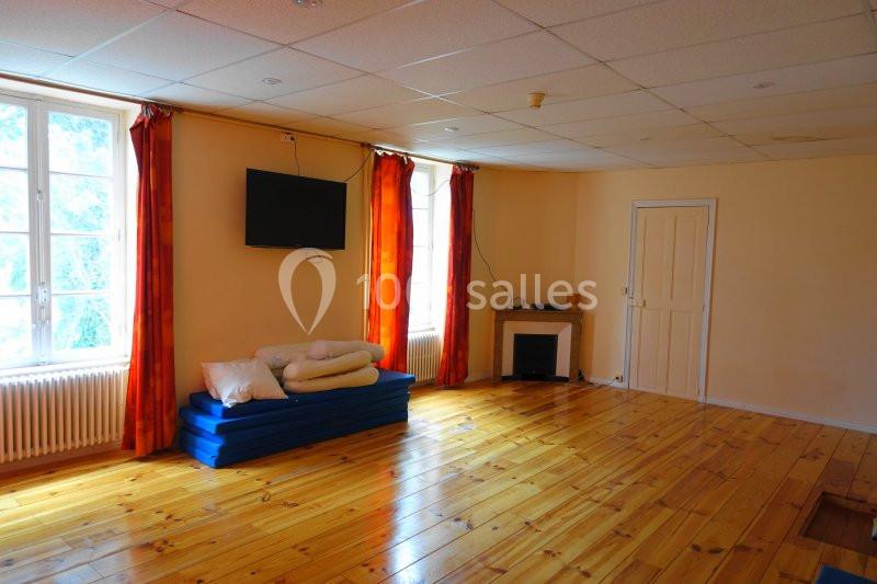 Salle lumineuse avec parquet en bois, tapis de sol empilés, coussins, télévision murale et fenêtres avec rideaux rouges.