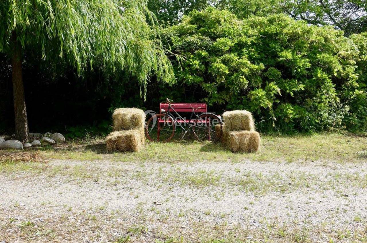 Banc rouge entouré de bottes de foin, placé sur un sol gravillonné devant des arbres et des buissons verdoyants.