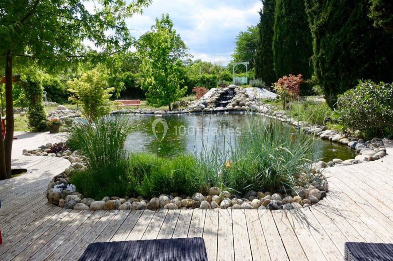 Étang entouré de végétation, bordé de pierres et d'une terrasse en bois, avec un petit pont visible au loin.