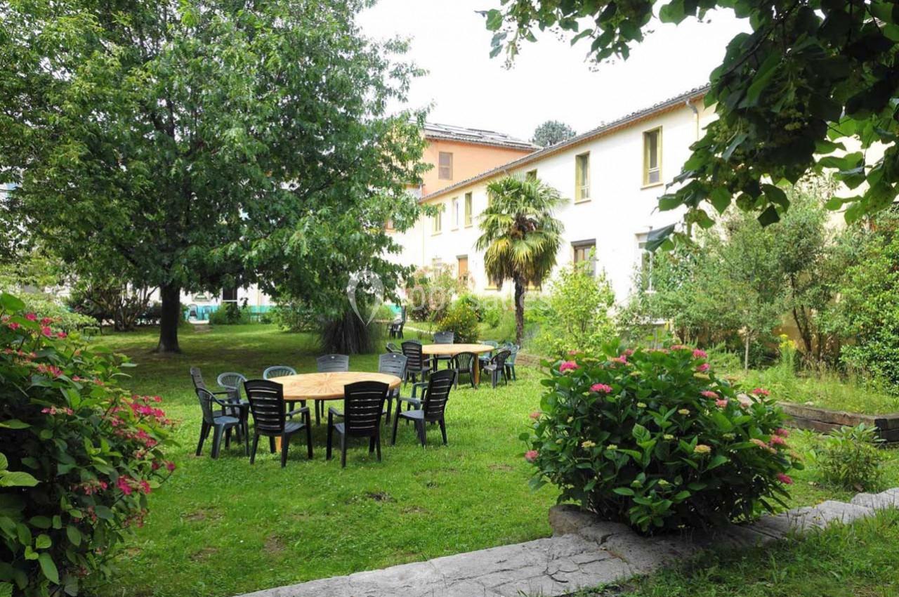 Jardin verdoyant avec tables et chaises en plastique noir, entouré d'arbres et de buissons fleuris devant un bâtiment clair.