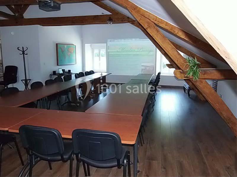 Salle de réunion sous combles avec poutres apparentes, tables en bois, chaises noires et écran de projection allumé.