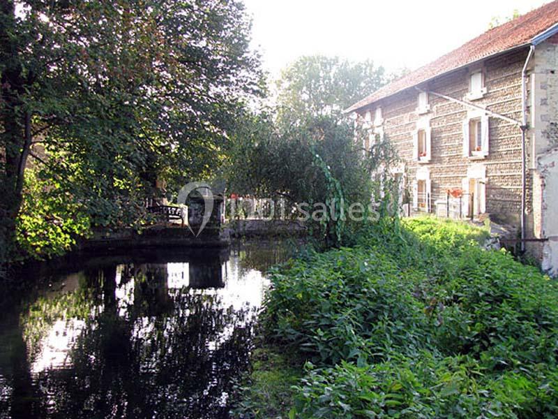 Vue d'un cours d'eau bordé de végétation, avec un bâtiment en pierre et des fenêtres visibles à droite.