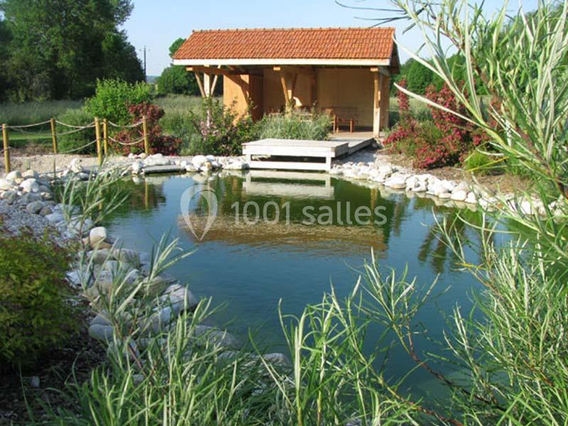 Étang paysager entouré de végétation et bordé de galets, avec un abri en bois au toit rouge en arrière-plan.