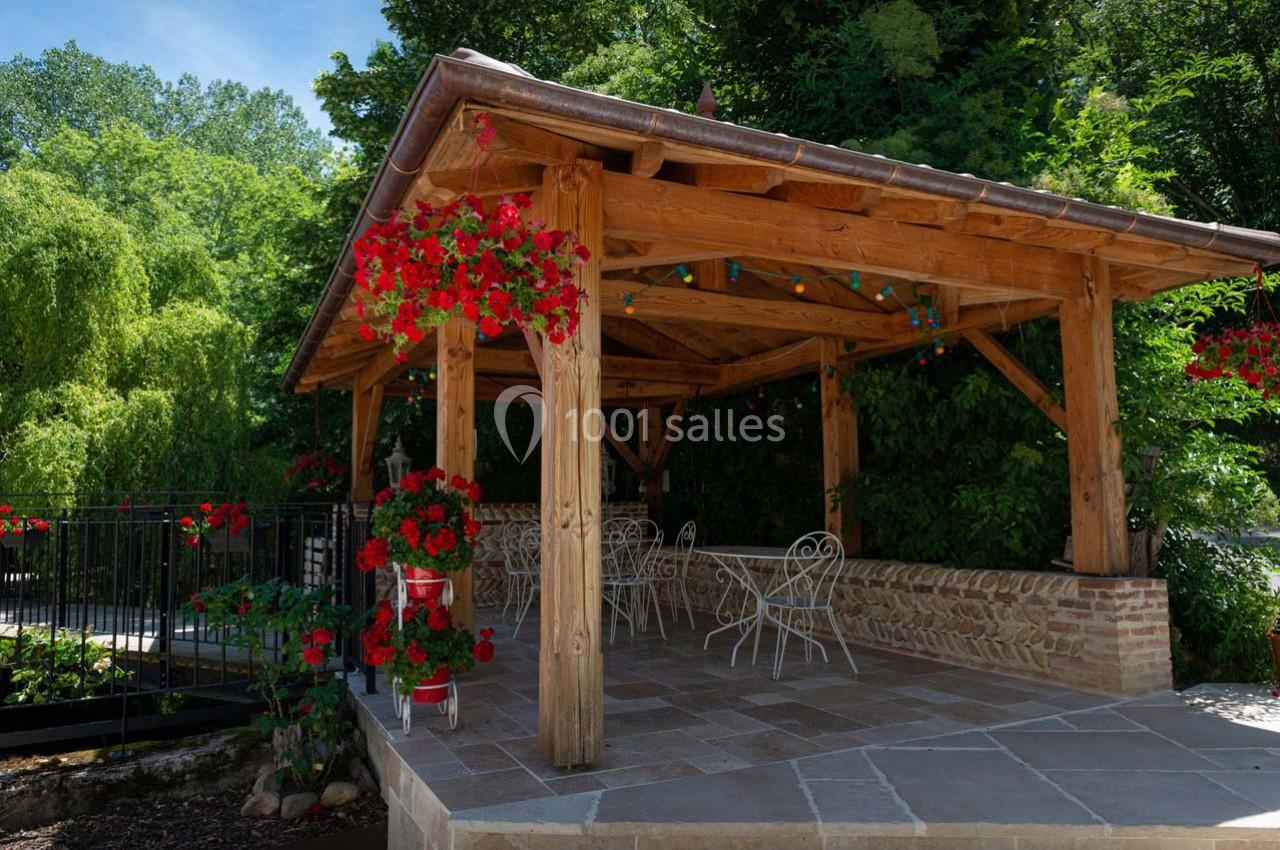 Pergola en bois avec des tables et chaises blanches, entourée de fleurs rouges et de verdure.