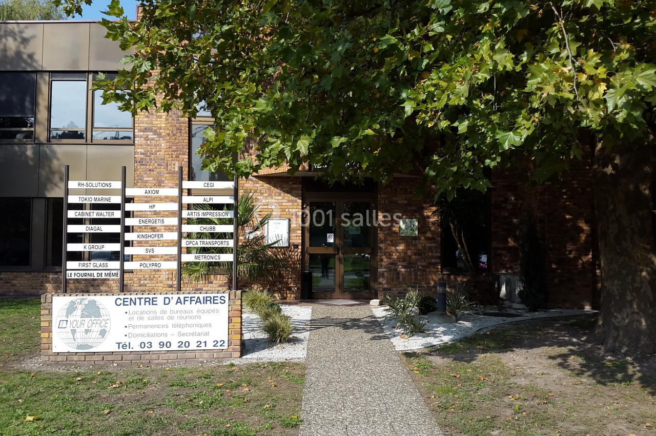 Façade d'un centre d'affaires en briques, avec une entrée bordée de végétation et des panneaux d'information.