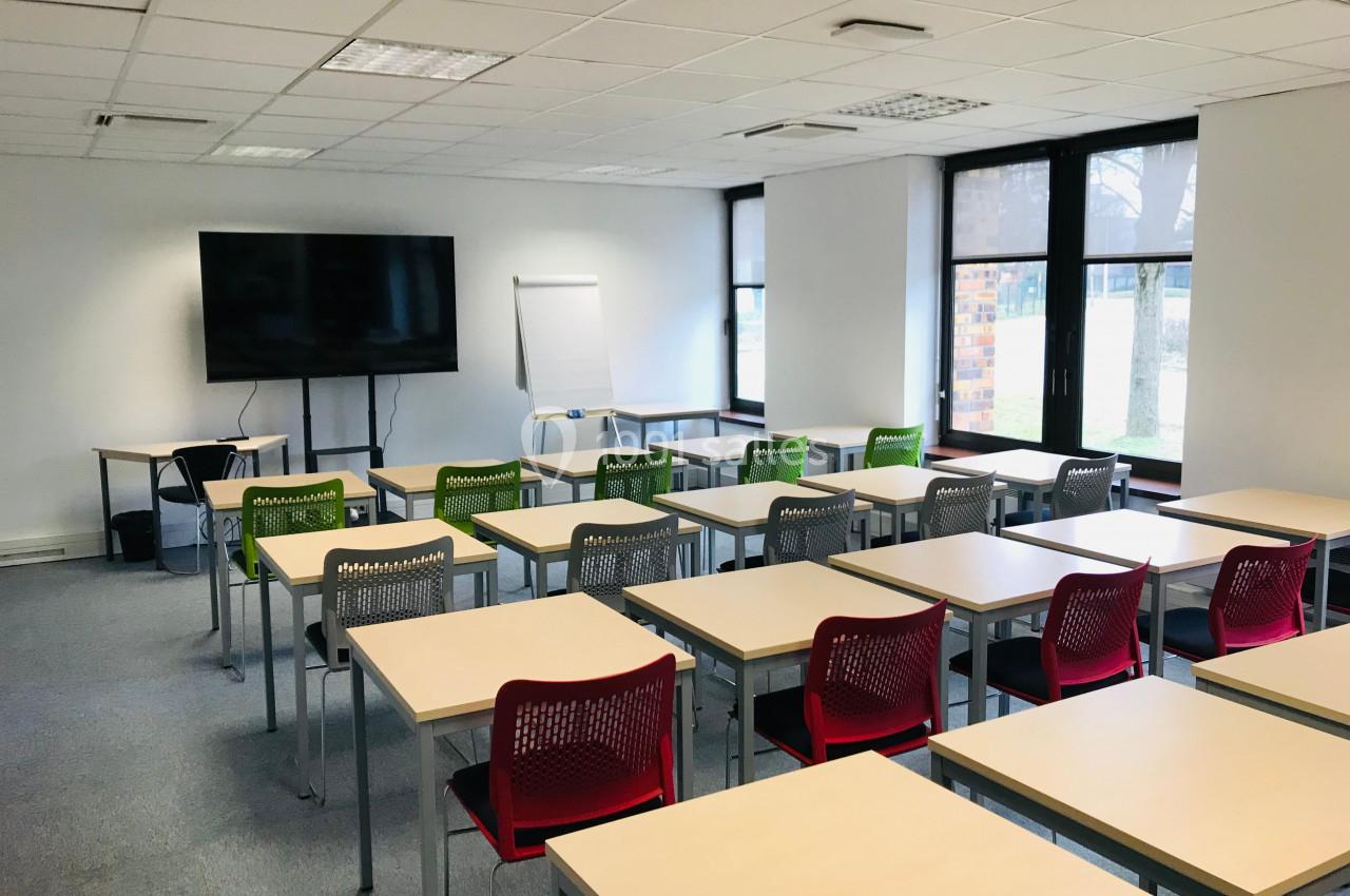 Salle de classe lumineuse avec des rangées de tables et chaises colorées, un écran et un tableau de présentation.