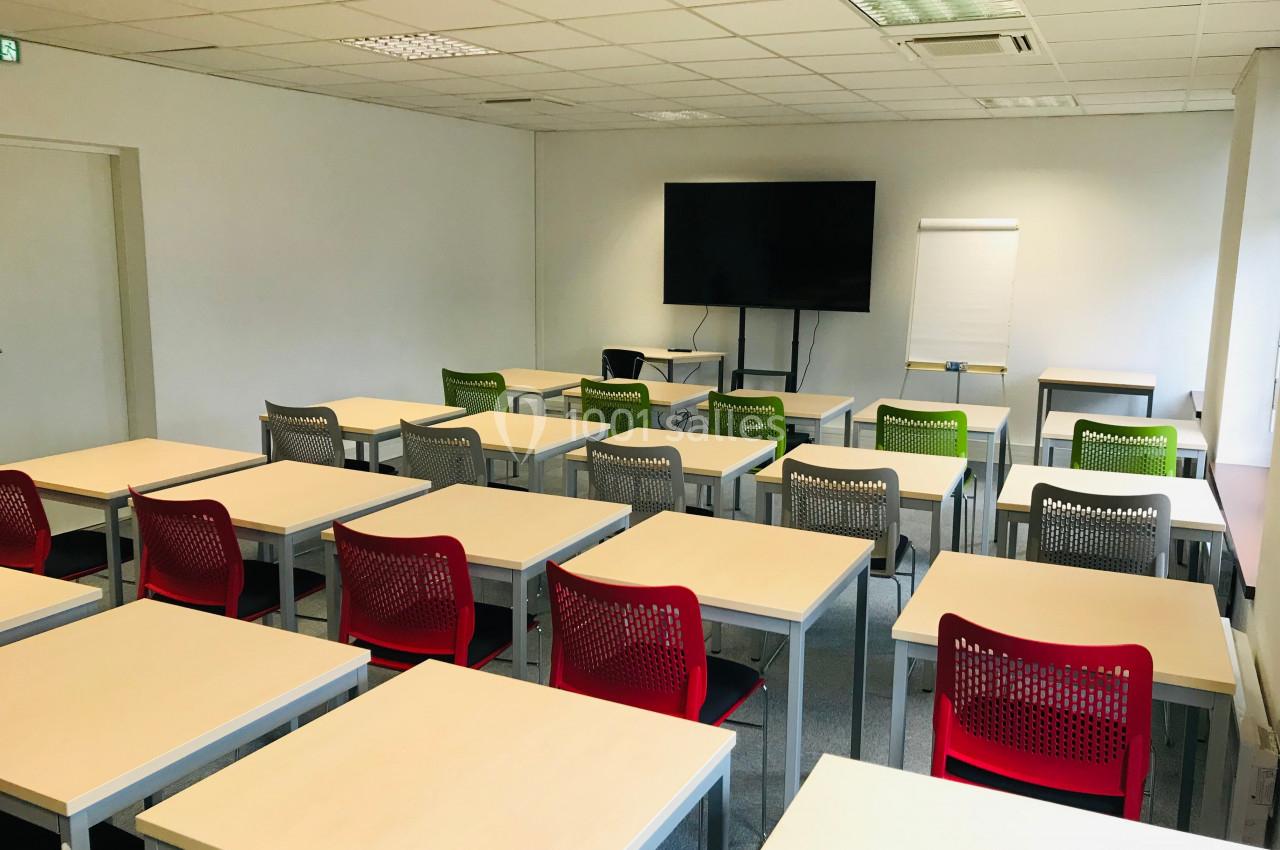 Salle de classe vide avec des tables, des chaises colorées, un tableau blanc, un écran et un paperboard.
