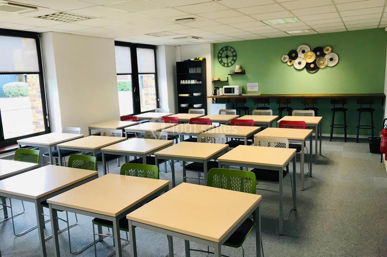 Salle de classe moderne avec tables et chaises alignées, mur vert décoré et coin kitchenette en arrière-plan.